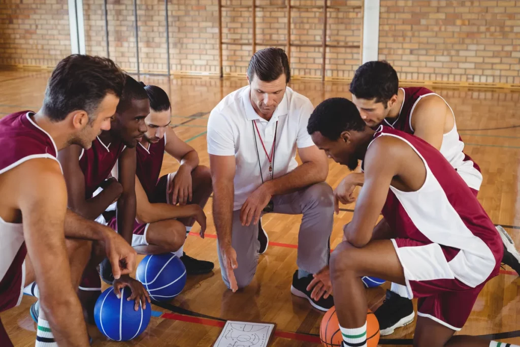 A coach in a white shirt huddled with a basketball team drawing up a play on the court, illustrating the manager-led daily huddle that drives dealership training results.